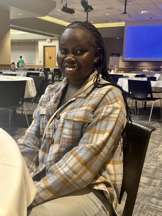 Image description: A photo of Agol Anei. She's wearing a plaid shirt and smiling toward the camera.